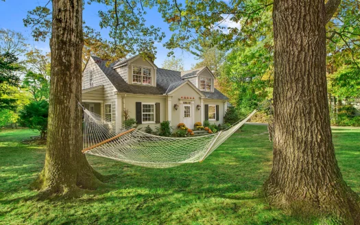 Cape Cod style house with fall mums and pumpkins on the front steps and a hammock between two oak trees
