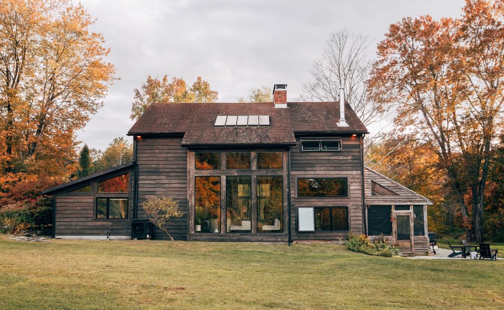Dark wood-sided country house with tall windows on a wide lawn with fall trees