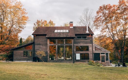 Dark wood-sided country house with tall windows on a wide lawn with fall trees