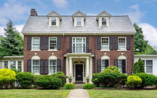 Brick center-hall colonial with black shutters, a slate roof and three dormers behind trimmed shrubs