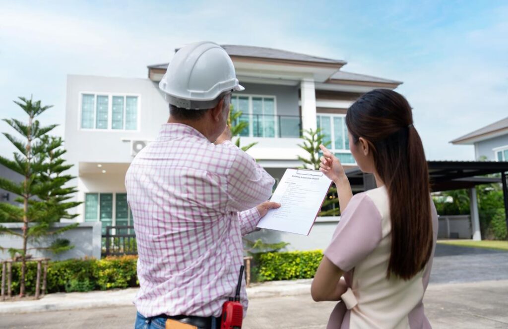 Home inspector in a hard hat reviewing a report with a buyer outside a house
