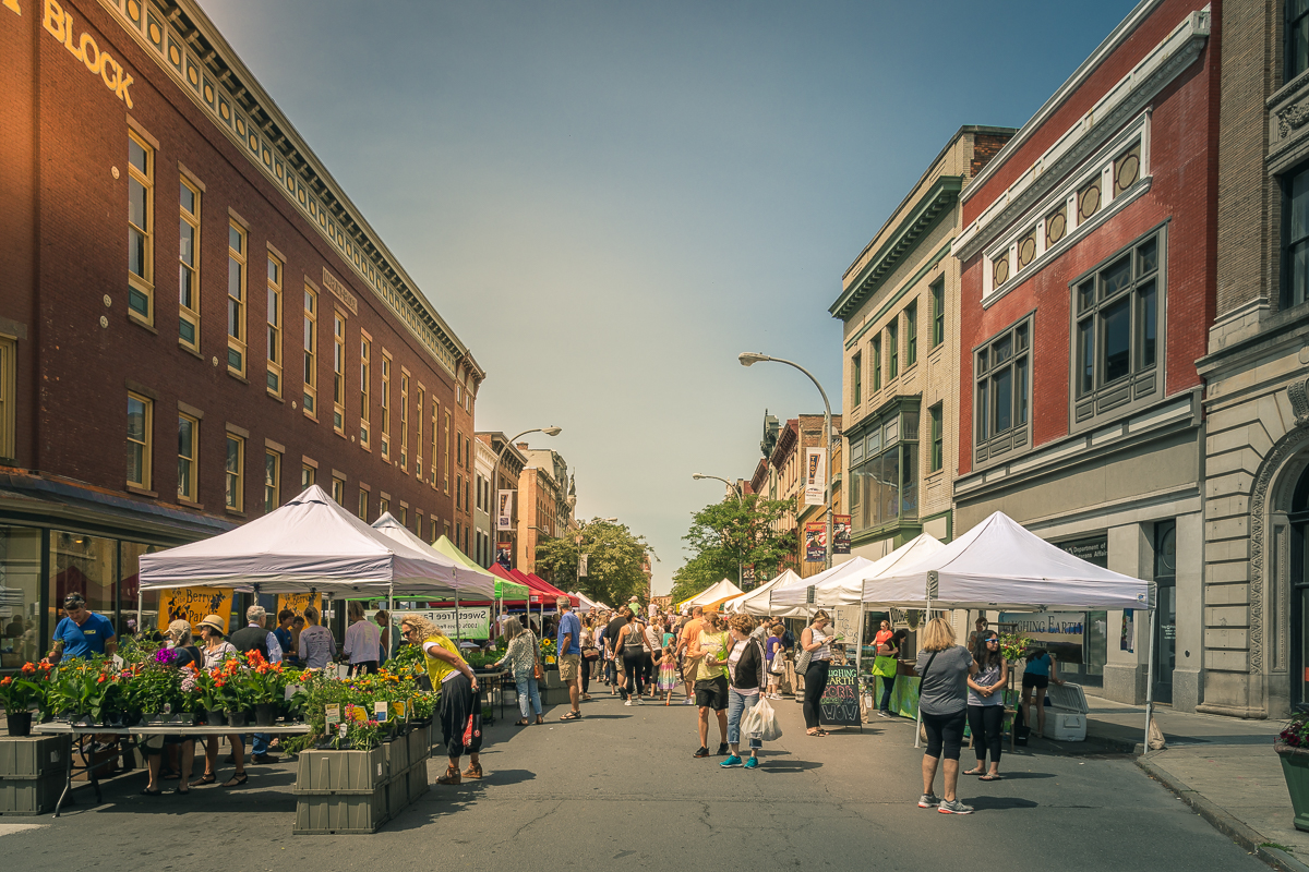 The Troy Waterfront Farmers Market on River Street on a Saturday morning — vendor tents, produce, and 19th-century downtown facades